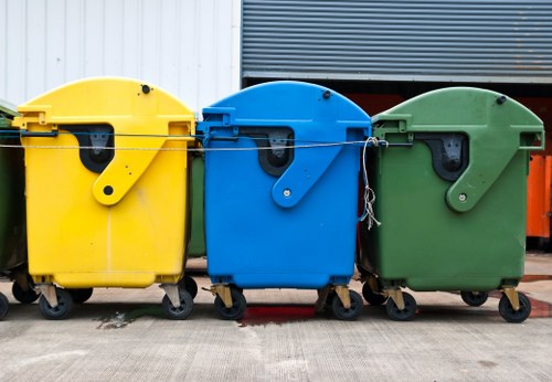 A row of three large, wheeled rubbish bins positioned side by side on a concrete surface, with the yellow bin on the left, the blue bin in the center, and the green bin on the right. Each bin has a sturdy, textured plastic body with a smooth, slightly curved lid that is closed, and black handles on the front for opening. The yellow bin appears to be for general waste, the blue bin for recycling, and the green bin for organic or garden waste, as is common in waste management services around Hornsey. They are situated outdoors, in front of a building with a partly visible metal facade and shutter, suggesting an industrial or commercial environment typical for waste collection in London or nearby areas. The scene is well-lit, with natural daylight casting subtle shadows beneath the bins, emphasizing their shapes and textures. The bins are aligned neatly and secured with black rims and small casters, with some minor wear visible on their surfaces, indicating regular use by Commercial Waste Removal Hornsey for efficient rubbish disposal and recycling collection services in the local area.