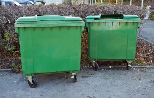Two green wheeled rubbish bins positioned on a paved outdoor surface, with the larger bin on the left and the smaller bin on the right, both featuring closed, hinged lids and visible wheels at the base for mobility. The bins are made of durable plastic with smooth, slightly matte surfaces and are placed side by side, with a small gap between them. In the background, there is a hedge with sparse, brownish foliage and a fence, indicating an outdoor setting possibly near a residential or commercial property in Hornsey. The scene appears well-lit, with natural daylight, emphasizing the clean, solid construction of the bins which are commonly used for waste collection and rubbish removal services. This image aligns with the services provided by Commercial Waste Removal Hornsey, focusing on waste management solutions in the local area, specifically within the context of rubbish disposal and collection.
