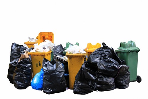 A collection of various rubbish bins and trash bags positioned outdoors on a paved surface, with a cluster of yellow, green, and black wheelie bins, some with their lids open or closed. The black plastic trash bags are piled around the bins, some partially leaning against them, filled with household waste or debris, and tied at the top. A blue plastic bag is also visible among the black bags. The setting appears to be a designated waste disposal or collection area, with the bins and bags arranged closely together, likely for a rubbish removal service. The background is plain and white, with no other environmental features visible, highlighting the focus on the waste containers and bags. This scene typifies a rubbish collection site accessible for professional waste removal services, such as those offered by Commercial Waste Removal Hornsey, serving the local area.