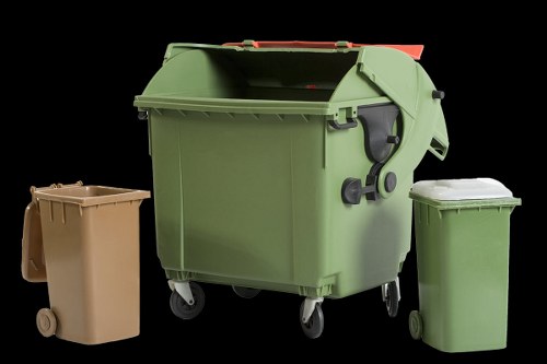 A large green wheeled waste storage bin with a closed lid positioned on a flat surface, accompanied by two smaller plastic waste bins—one beige and one green—placed nearby. The green bin has a textured surface, a black handle, and is situated outdoors, with a dark background highlighting its vibrant color. The beige bin is made of smooth plastic with small wheels, while the green bin has a smoother finish and is slightly taller. The scene appears to depict a setting related to waste collection or rubbish disposal, reflecting the services offered by Commercial Waste Removal Hornsey for local rubbish management. The objects are arranged in a manner typical for ready collection, emphasizing the practical aspect of waste removal services in the Hornsey area.