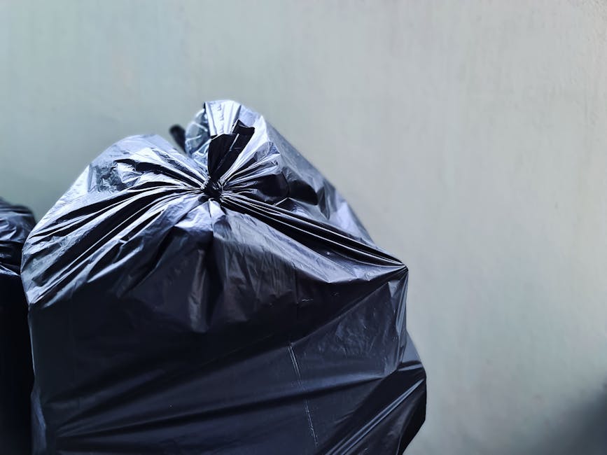Close-up of a tightly knotted black plastic rubbish bag made from glossy, flexible material, placed on a surface with additional similar bags partially visible on the left. The bag's surface exhibits