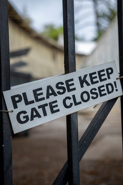 A close-up view of a white rectangular sign with bold black lettering that reads 'Please Keep Gate Closed!', attached to a black metal gate with vertical and diagonal bars. The gate appears to be part